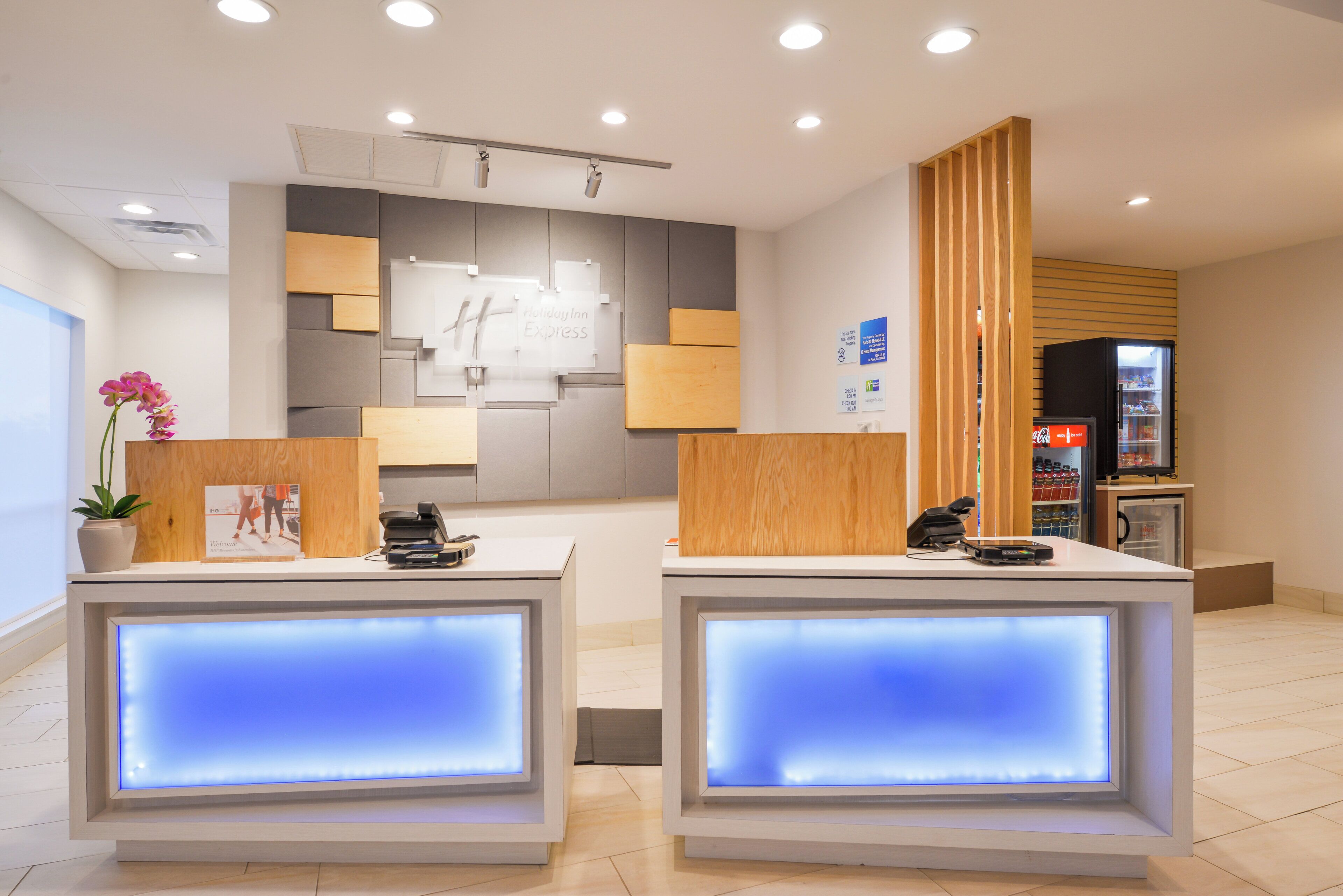 Uma área de recepção de hotel com dois balcões, uma planta e um refrigerador abastecido com bebidas.