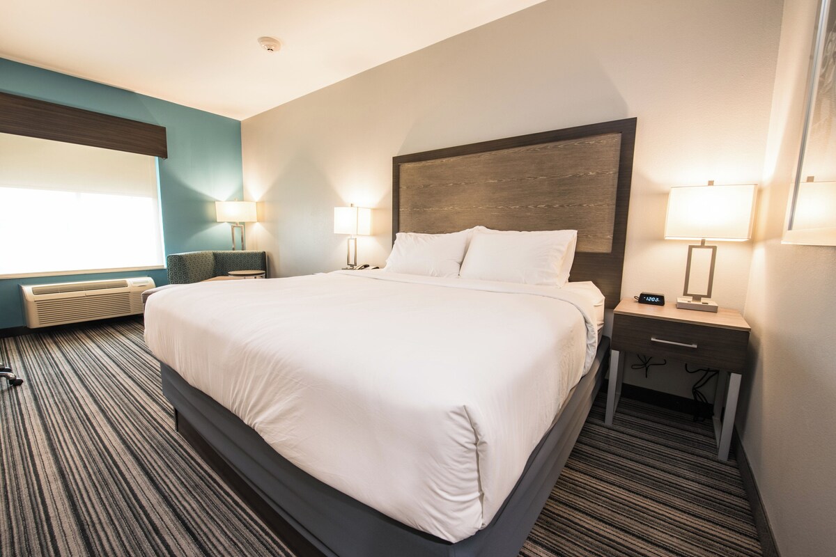 A hotel room with a large bed, two bedside tables with lamps, a window with blinds, and a carpeted floor.