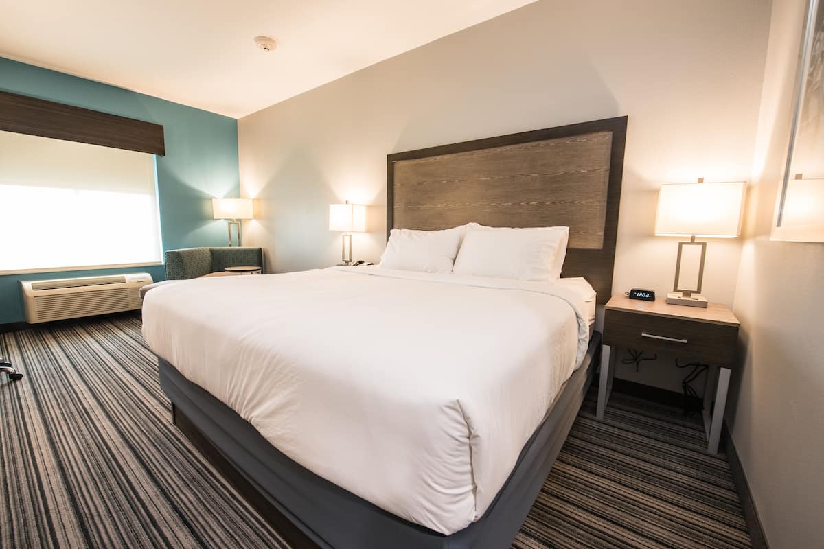 A hotel room with a large bed, two bedside tables with lamps, a window with blinds, and a carpeted floor.