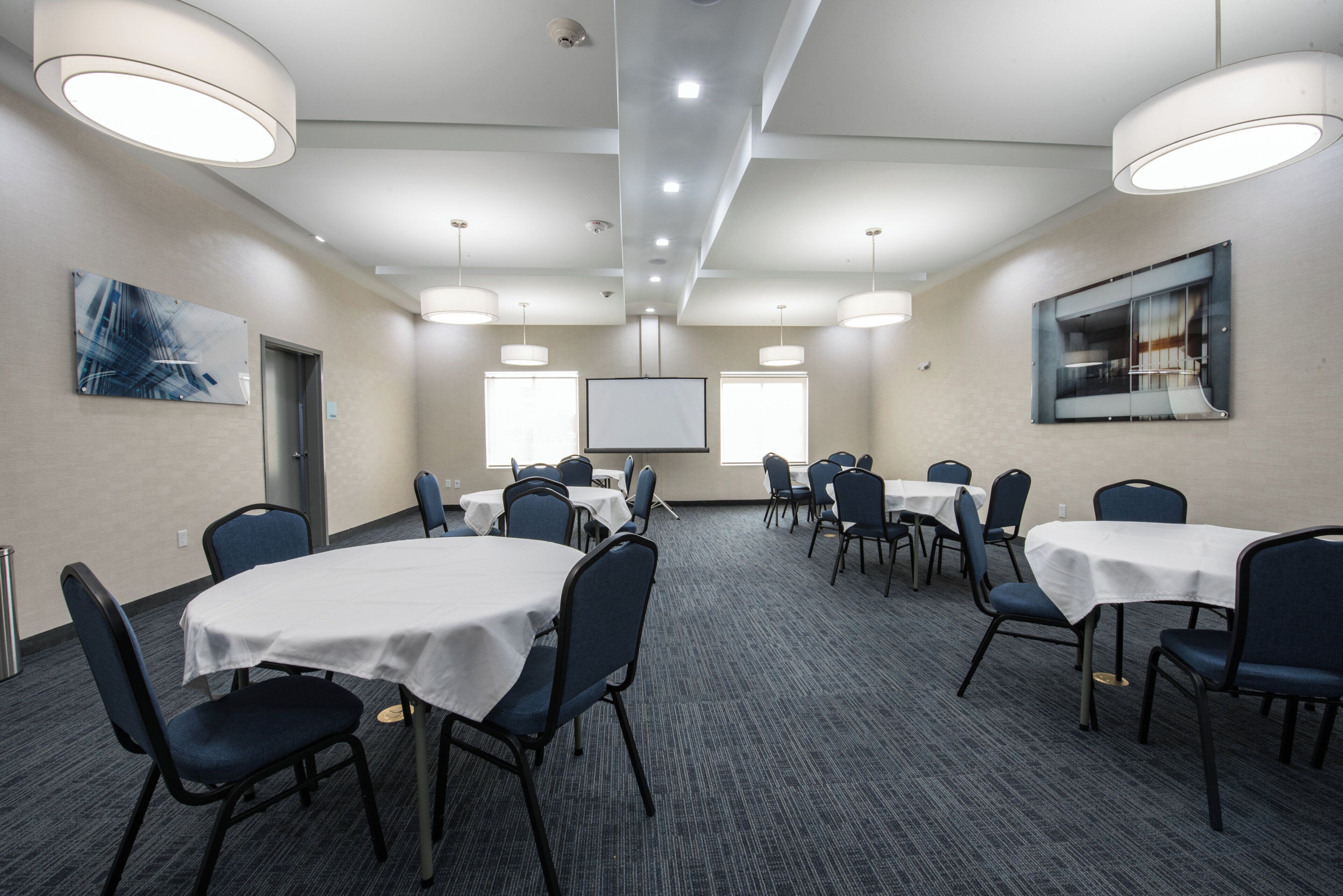 A conference room with multiple tables and chairs, a projector screen, and a large painting on the wall.
