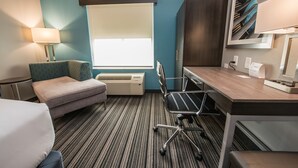 Et hotelværelse med seng, skrivebord, stol, lampe, et vindue med rullegardiner, en aircondition og et billede hængt på væggen.