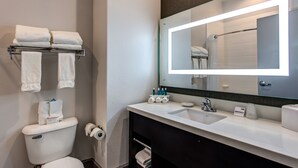 Un baño moderno con un lavabo blanco, un espejo grande con iluminación integrada y un inodoro. Hay toallas y artículos de aseo en la encimera del lavabo.