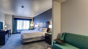 Una habitación de hotel con cama, mesitas de noche, escritorio, sofá verde y ventana con cortinas.