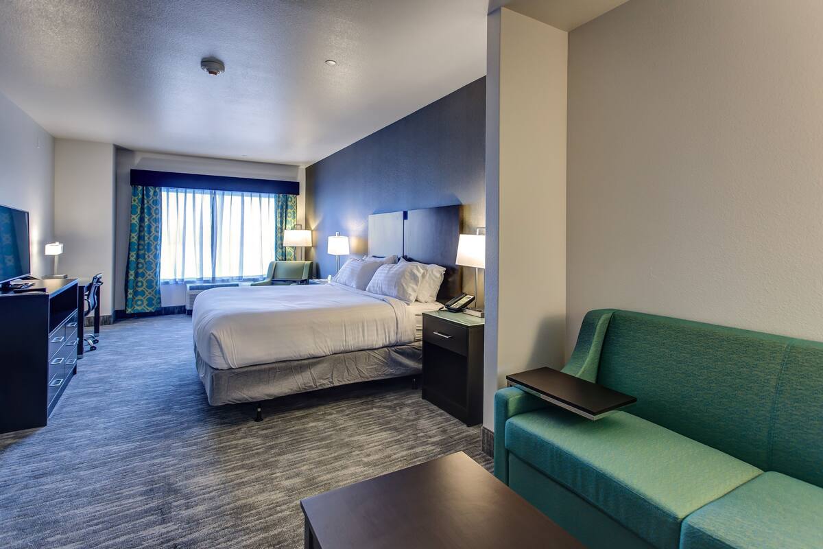 A hotel room with a bed, bedside tables, a desk, a green sofa, and a window with curtains.