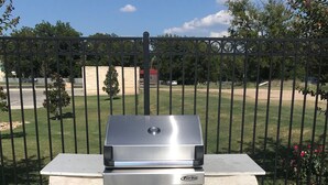 Una parrilla a gas con pantalla digital y cuatro quemadores, sobre una base de piedra, frente a una cerca metálica y follaje.