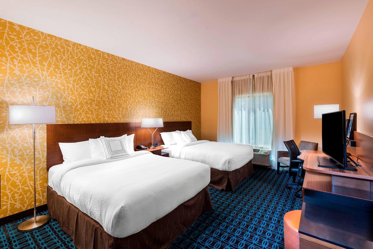 A hotel room with two beds, a desk, a TV, and a large window with curtains.