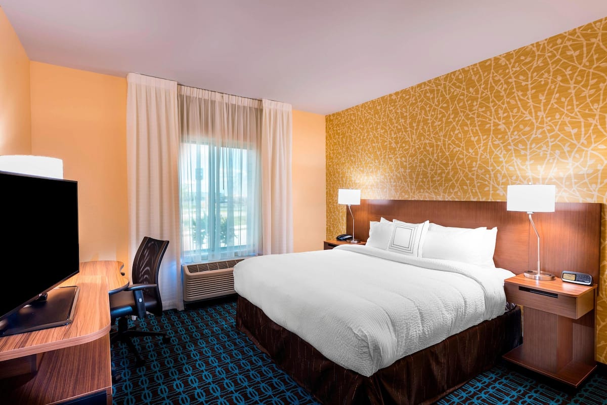 A hotel room with a large bed, a desk with a flat-screen TV, a chair, a nightstand with lamps, and a patterned wall.