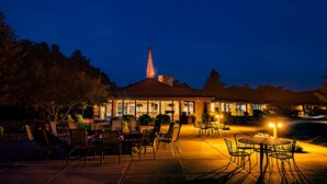 Un patio al aire libre con mesas y sillas, un edificio con una torre destacada y una entrada bien iluminada.