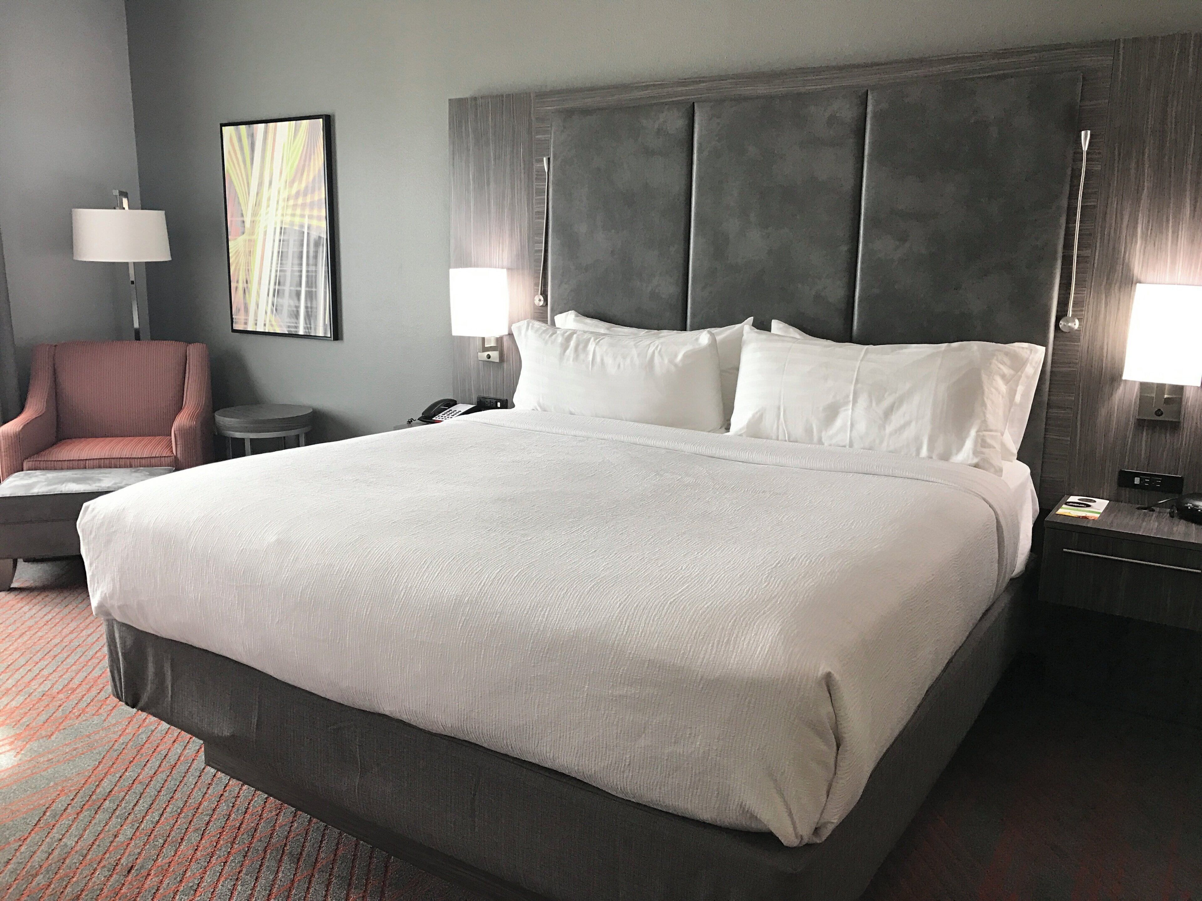 A large bed with a grey headboard, two bedside lamps, a telephone, and a chair in a hotel room.