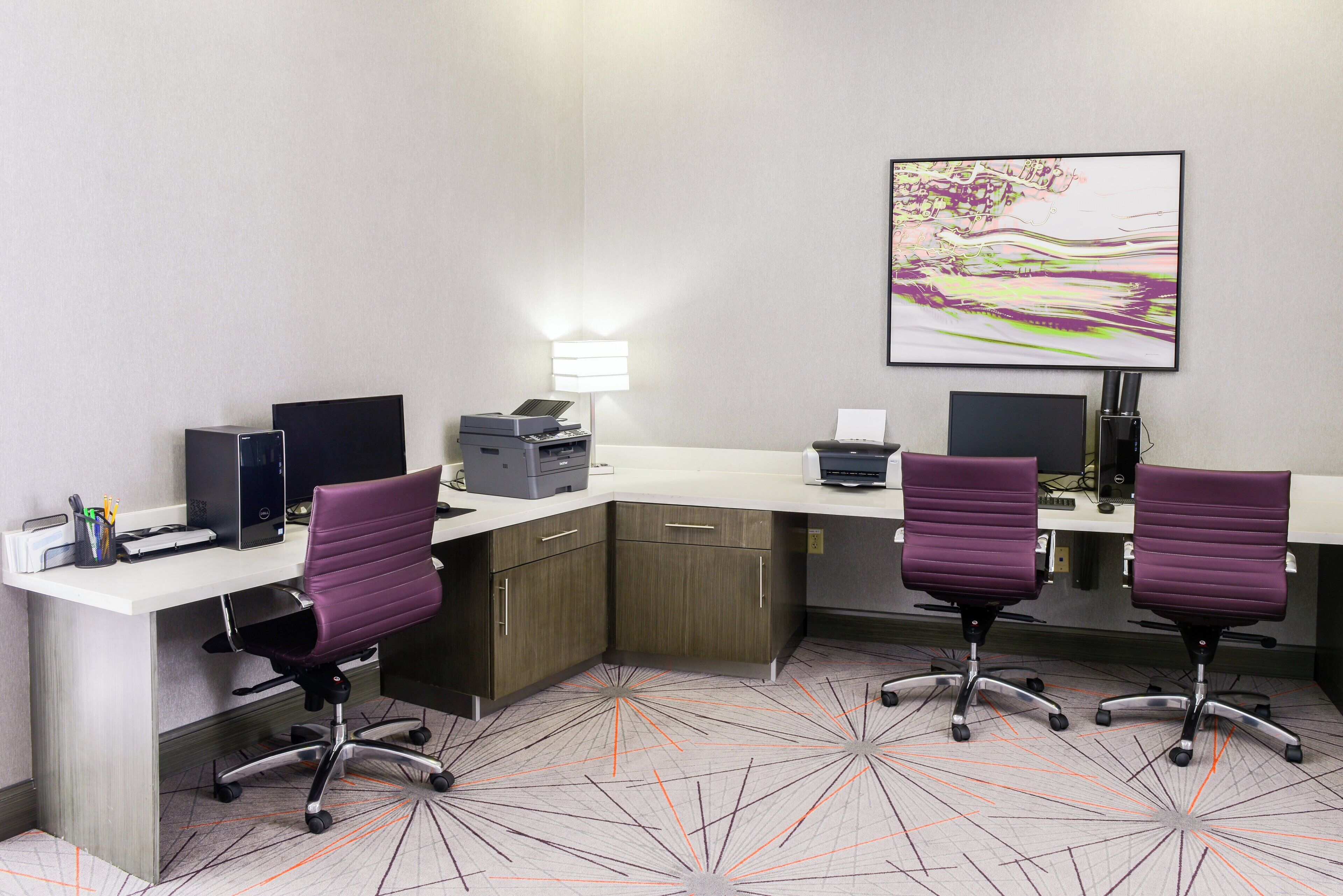 A modern office space with two desks, chairs, computers, printers, and a large abstract painting on the wall.