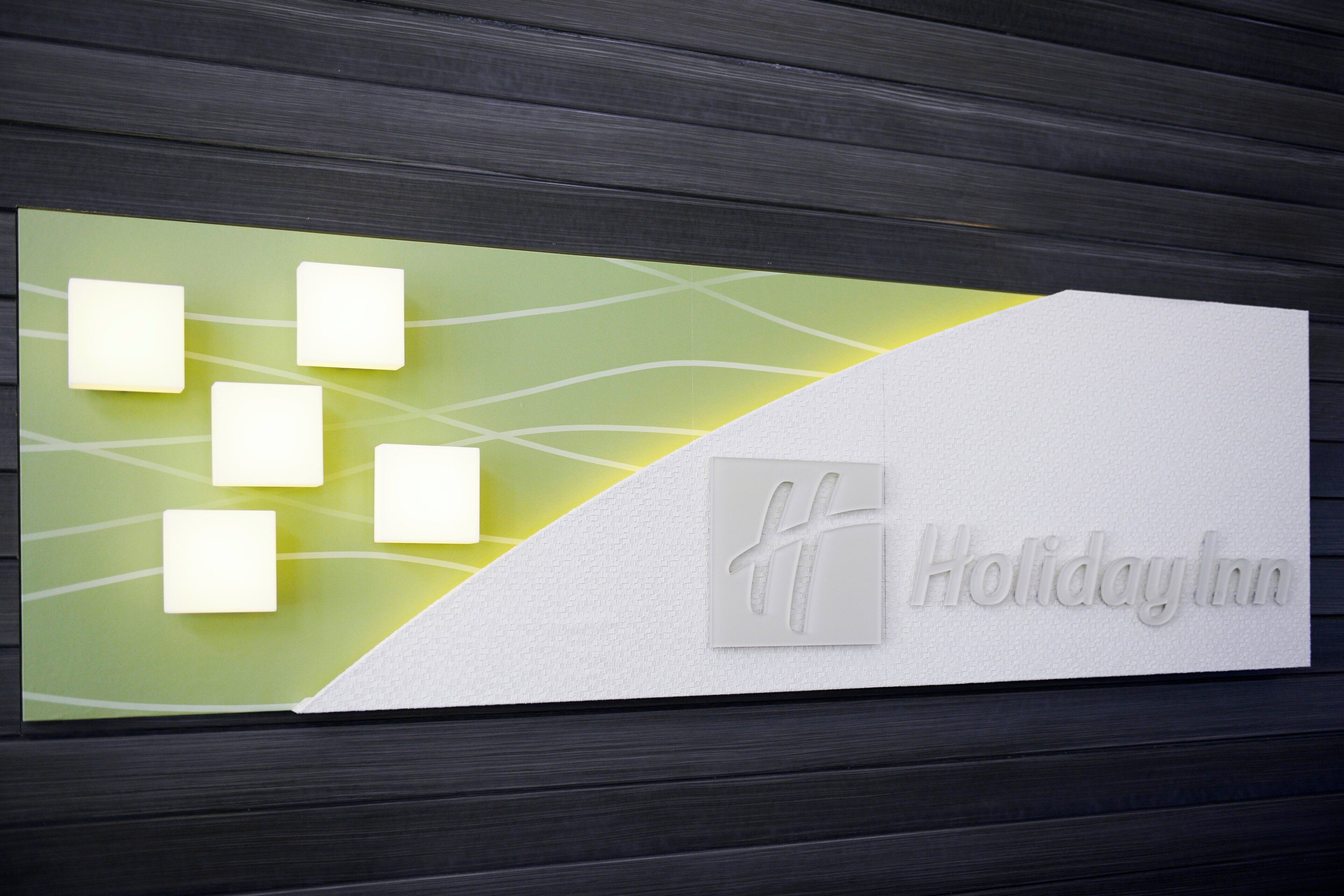 En skylt med texten ”Holiday Inn” och en logotyp, placerad på en vägg med en mörk träpanel som bakgrund.