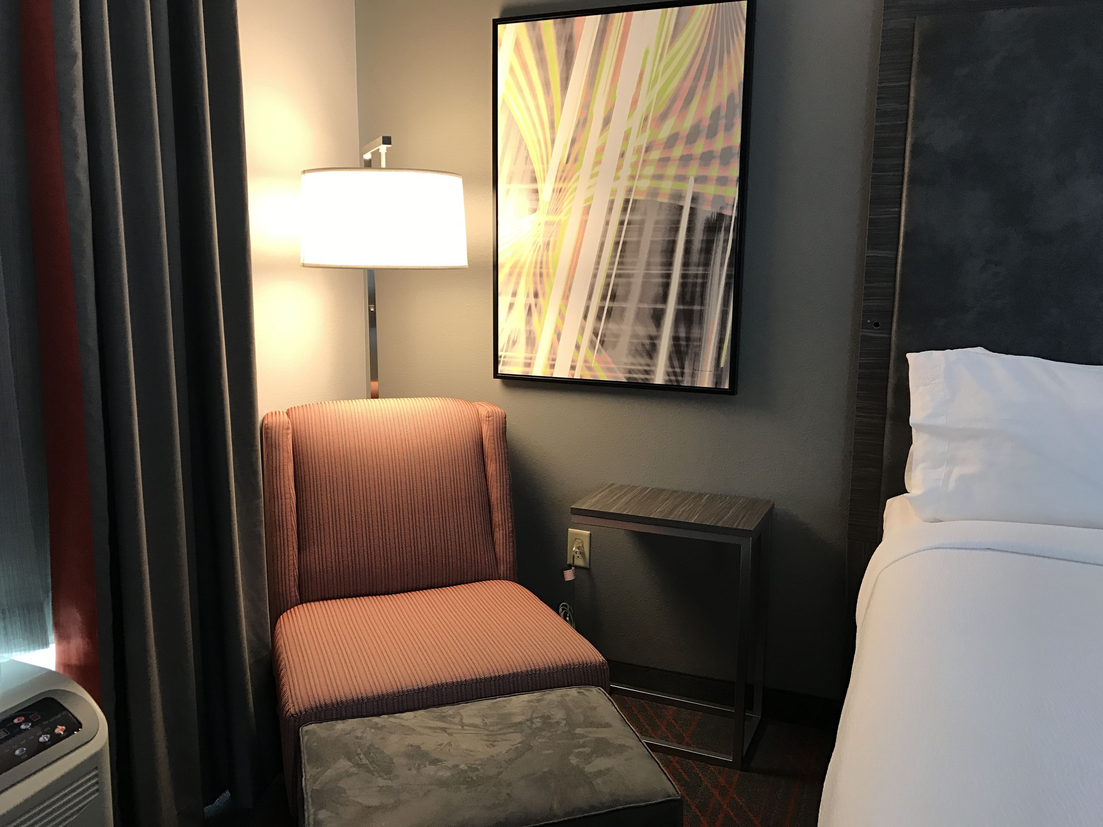 A hotel room with a bed, a chair, a small table, a lamp, and a wall-mounted artwork.