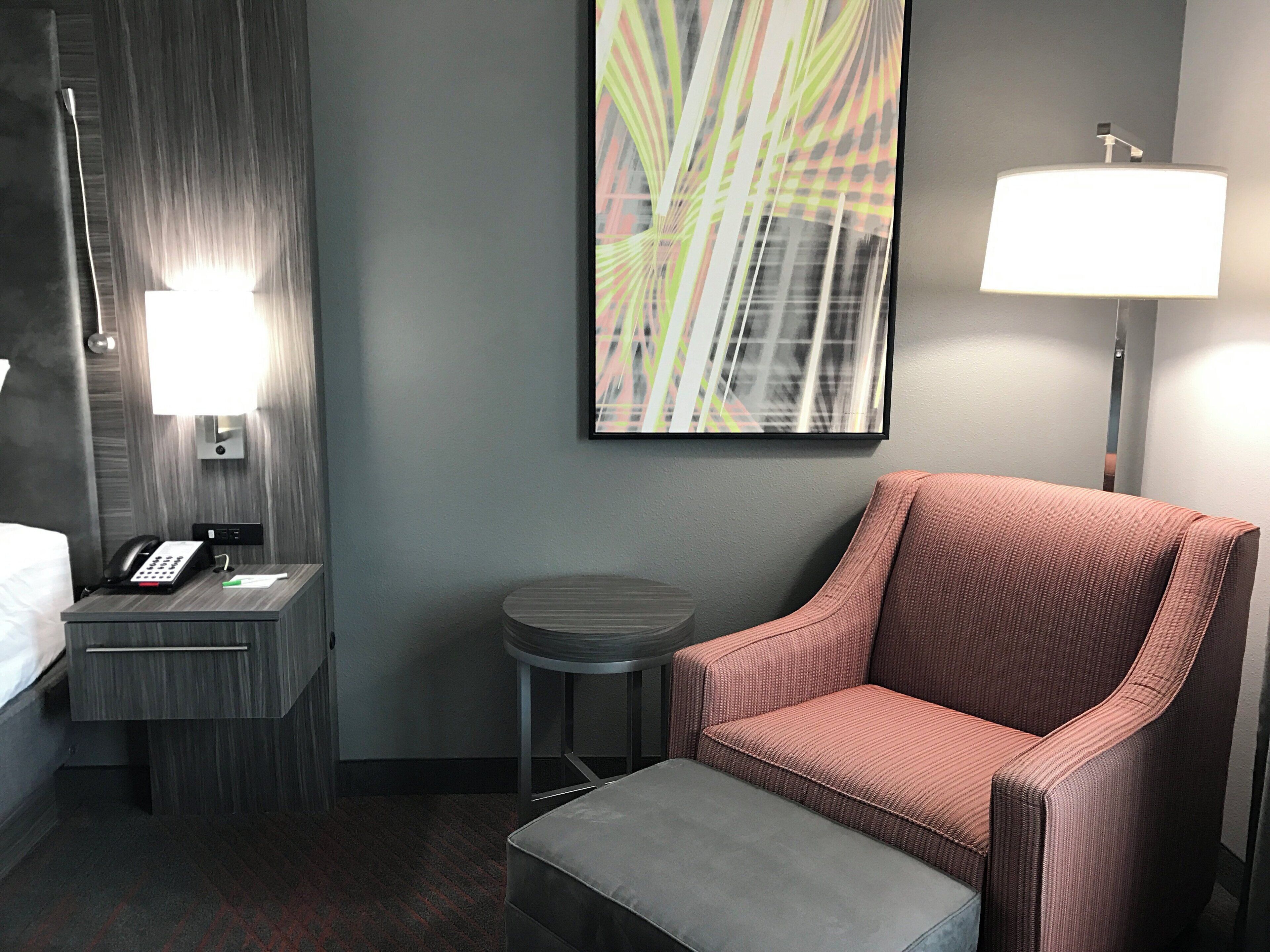 A hotel room with a bed, a desk with a phone, a chair, a small table, a lamp, and a painting on the wall.