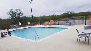 Une piscine entourée d’une clôture et d’arbres, avec des chaises longues et une table à proximité.