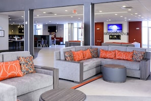 A modern lounge area with grey sofas and orange patterned cushions, a round coffee table, and a flat-screen TV mounted on the wall.