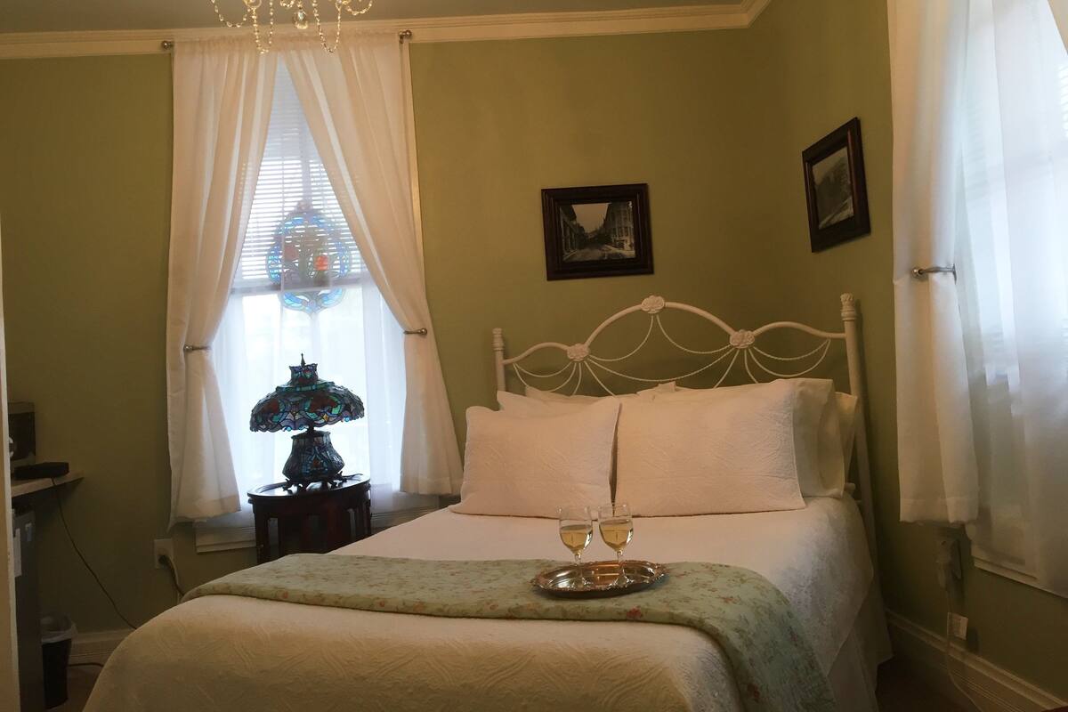 A bedroom with a bed, a chandelier, a window with curtains, and a small table with two glasses.