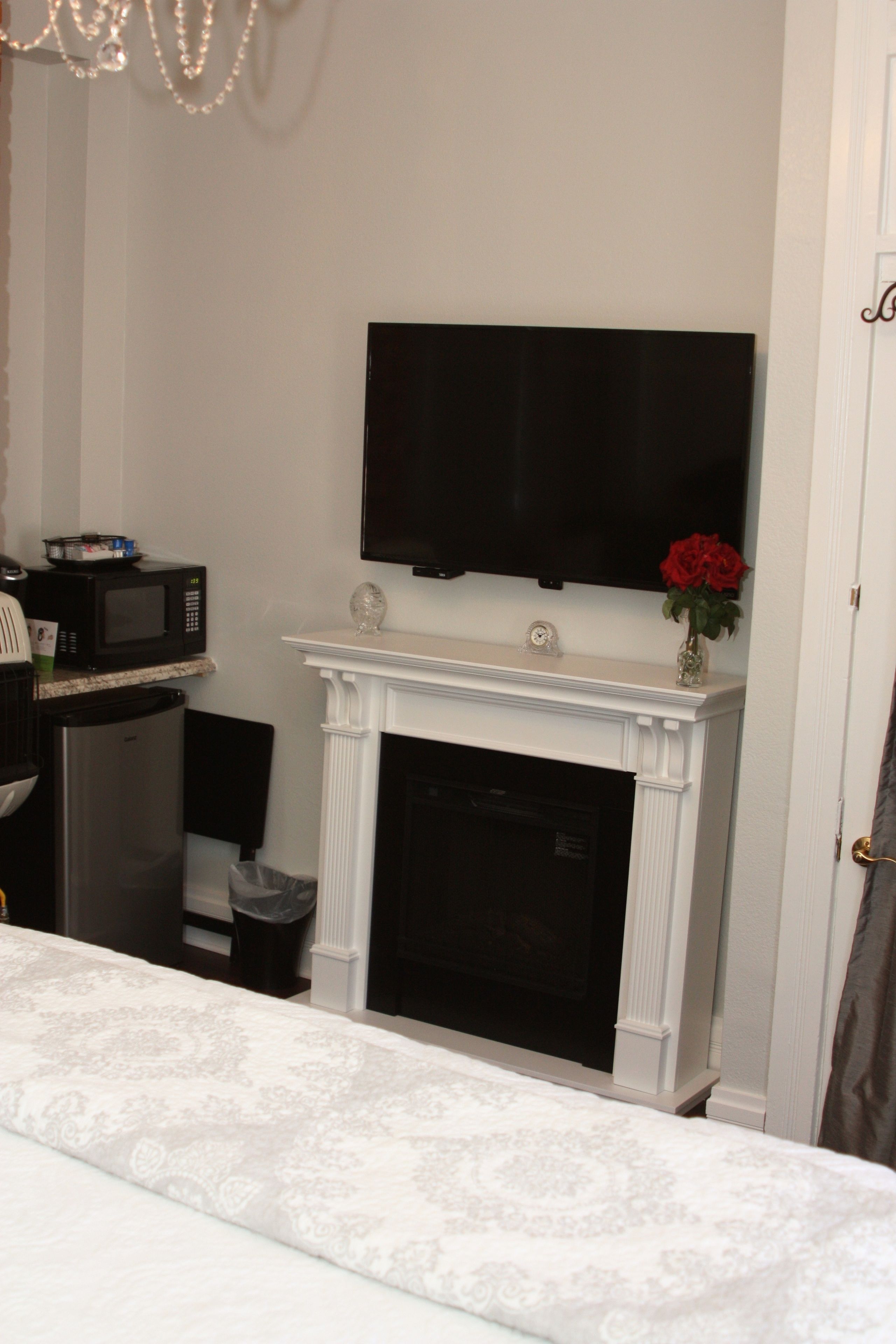 A room with a white fireplace, a flat-screen TV mounted above it, a microwave on the counter, and a trash can beside the fireplace.