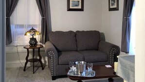 A living room with a grey sofa, a small side table with a lamp, a tray with glasses and a candle, and a stained glass chandelier. - Elmwood House (Eureka Springs)