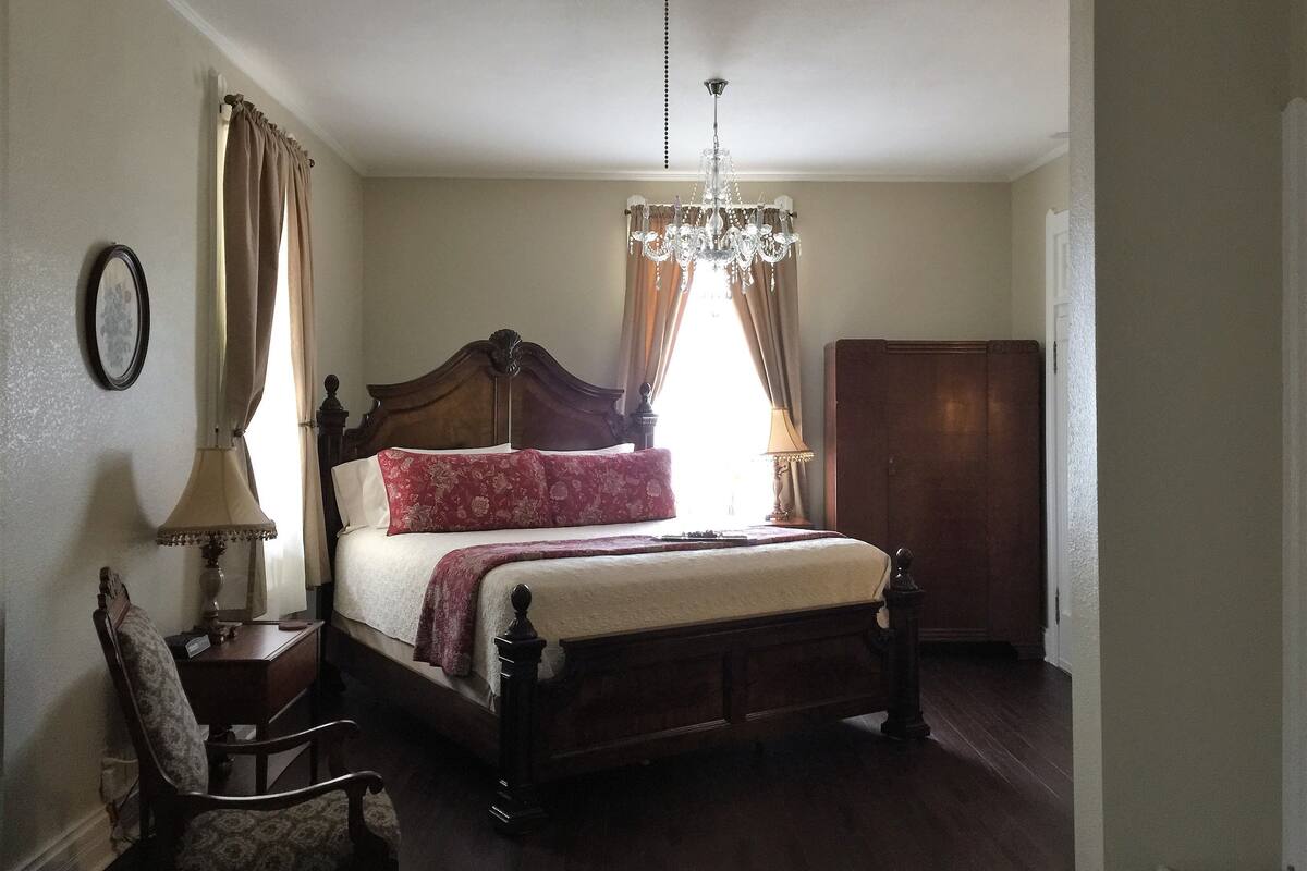A bedroom with a large bed, a wooden wardrobe, a chair, a bedside table with a lamp, and a chandelier.