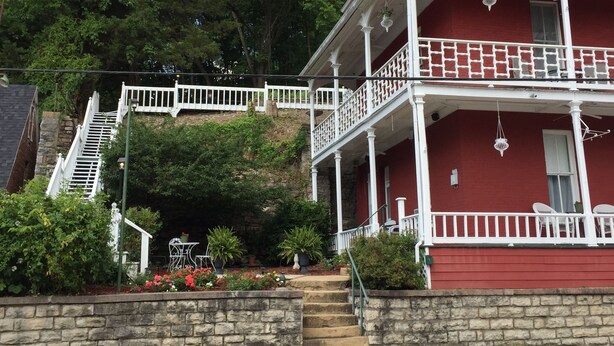 A two-story building with a red exterior, white balcony railings, and a stone staircase leading to the entrance. - Elmwood House (Eureka Springs)