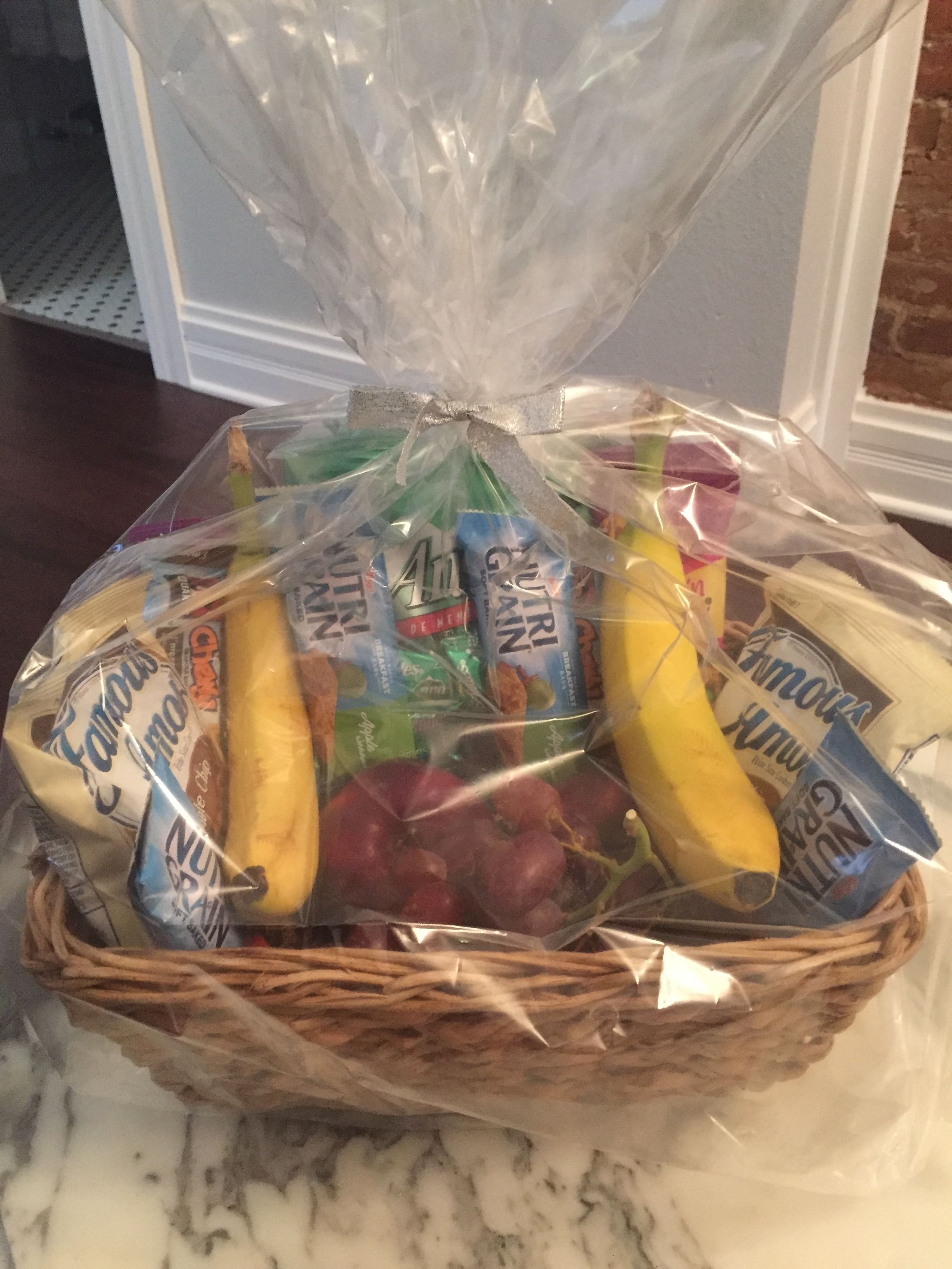 A wicker basket filled with various food items, including bananas, grapes, and packaged snacks, wrapped in clear plastic.