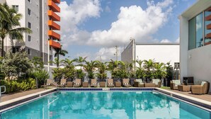 Une piscine entourée de bâtiments, avec des chaises longues et des plantes en pot, sous un ciel dégagé.