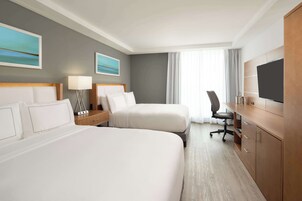 A hotel room with two beds, a desk, a chair, a TV, and artwork on the walls.