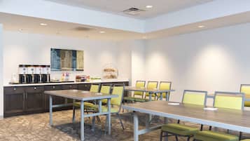 Un salón de conferencias con una larga mesa y sillas, una kitchenette con cafetera y un cuadro en la pared.