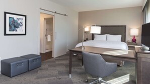 A hotel room with a bed, desk, chair, and a small seating area. There is a door leading to another room and a framed picture on the wall. - Hampton Inn & Suites Chicago Schaumburg (Schaumburg)