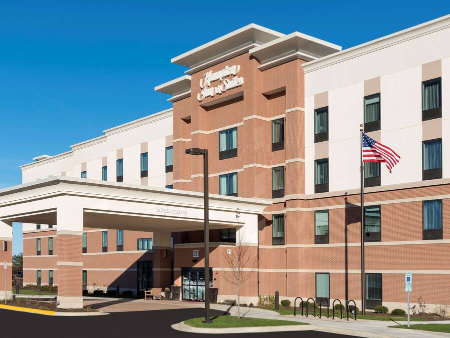 Un bâtiment de l’hôtel Hampton Inn & Suites, avec une entrée bien visible, un drapeau américain et une pancarte « Bienvenue ».