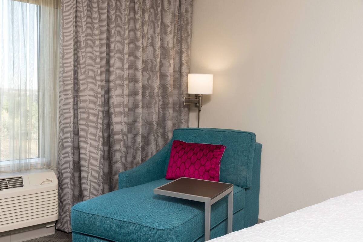 Un fauteuil turquoise, accompagné d’une petite table et d’un coussin rose, d’une lampe blanche et d’une fenêtre ornée de rideaux légers.