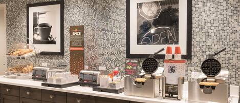 A breakfast bar with various food items and appliances, including a waffle maker and a coffee machine.
