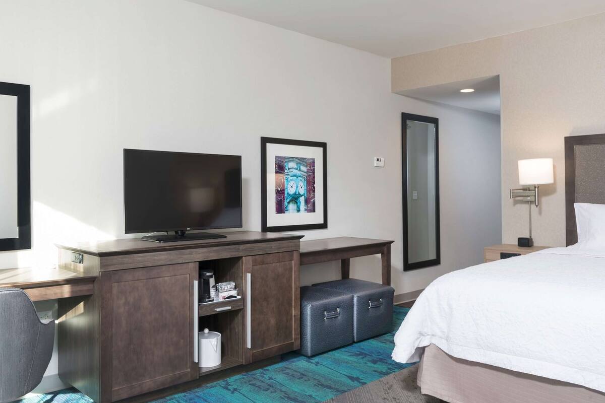 Une chambre d’hôtel comprenant un lit, un bureau avec un téléviseur à écran plat, une petite armoire, un miroir et une table de chevet avec une lampe.