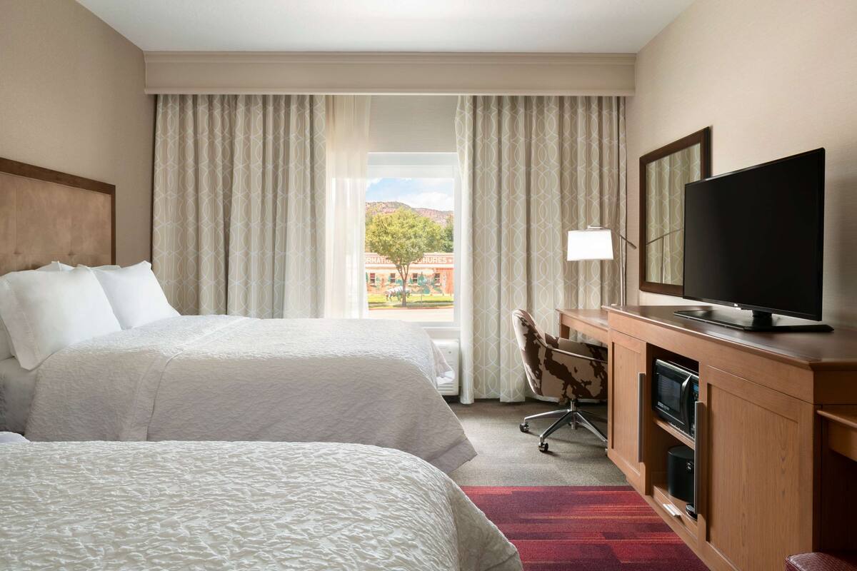 A hotel room with two beds, a TV, a desk, and a view of a building and trees outside.