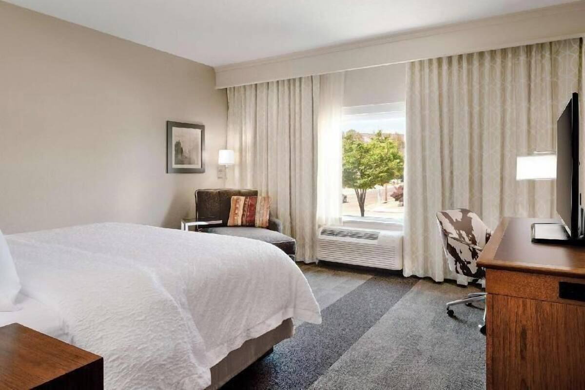 Une chambre d’hôtel avec un lit, un bureau, une chaise, une fenêtre avec des rideaux et un tableau au mur.