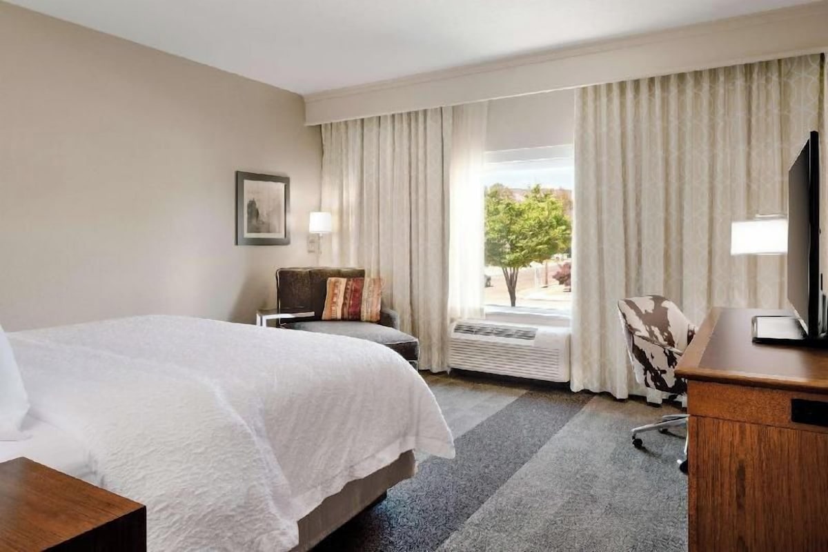 A hotel room with a bed, a desk, a chair, a window with curtains, and a picture on the wall.