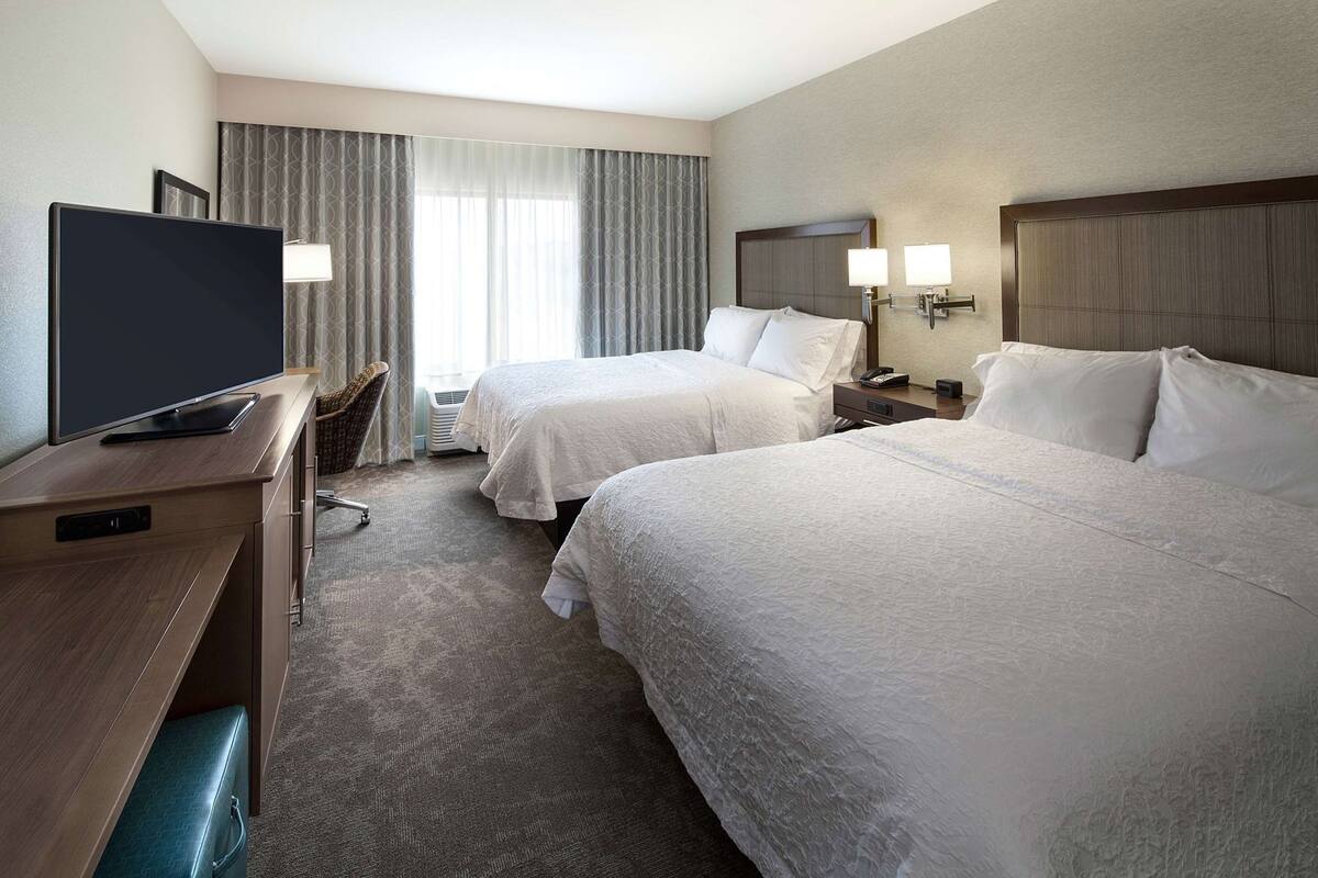 Quarto de hotel com duas camas, uma mesa de escritório com televisão, uma cadeira e uma mesa de cabeceira com luminárias.