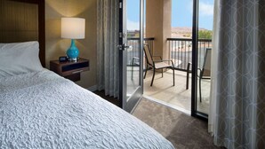 A hotel room with a bed, a nightstand with a lamp, a sliding glass door to a balcony with a chair and table.