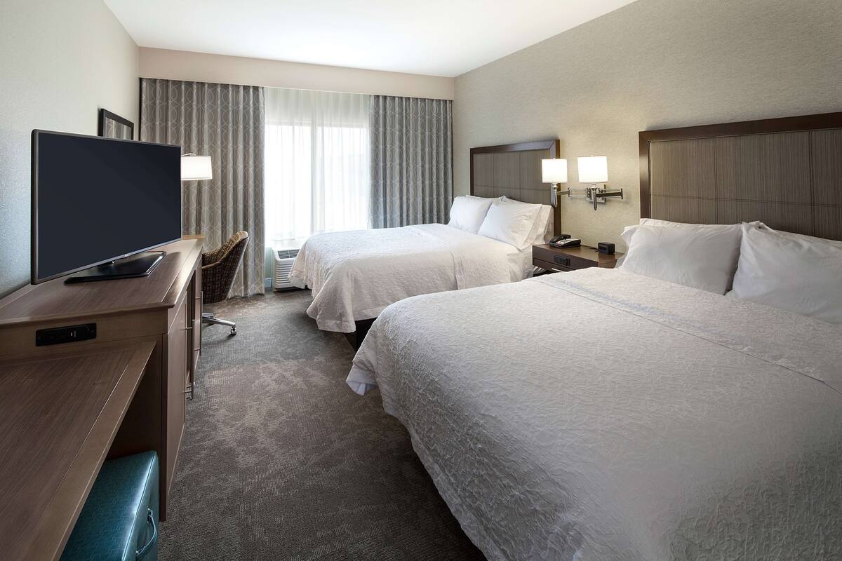 Quarto de hotel com duas camas, uma escrivaninha com televisão, uma cadeira e uma mesa de cabeceira com abajures.