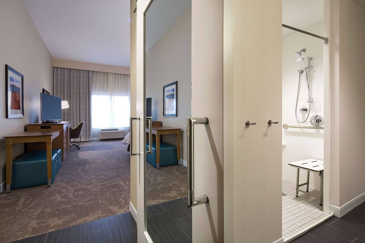 Quarto de hotel com uma mesa, cadeira e uma pequena mesa. É visível uma área de chuveiro através de uma porta aberta.