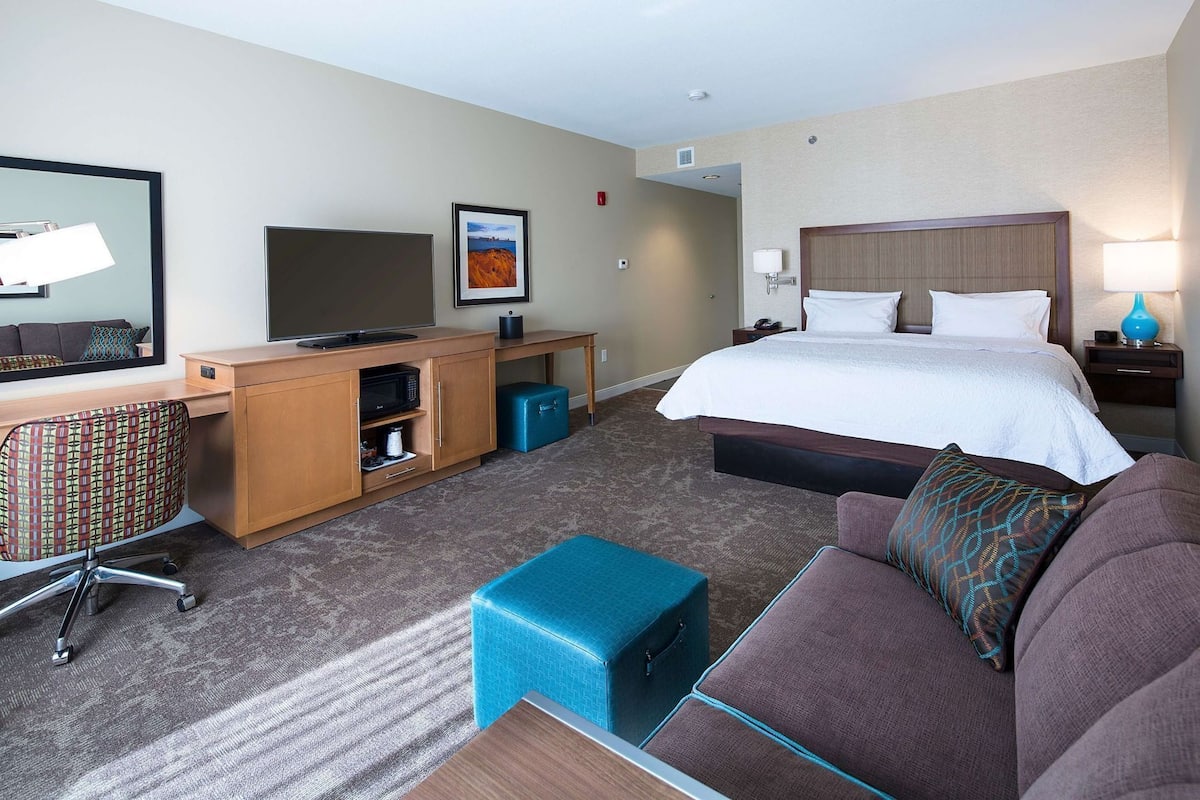 Quarto de hotel com cama, escrivaninha com computador, televisão, sofá e um pequeno banquinho azul.