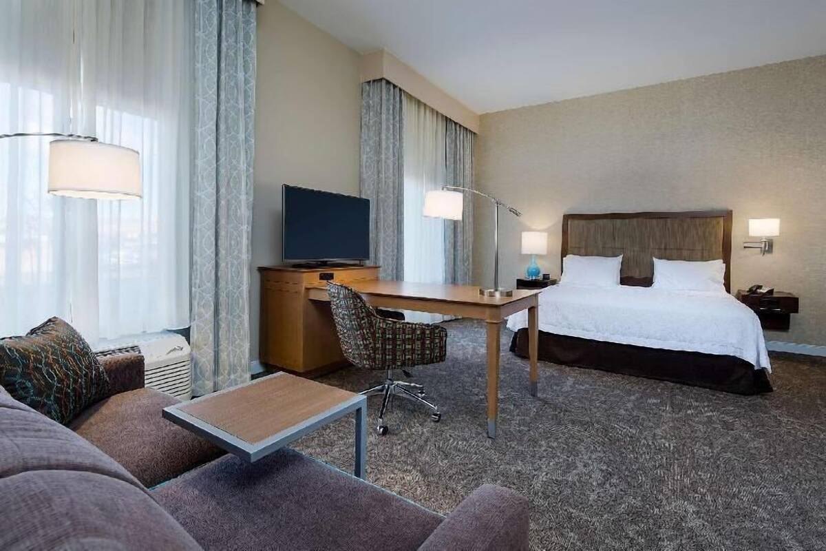 Quarto de hotel com cama, escrivaninha, cadeira, televisão e luminárias.
