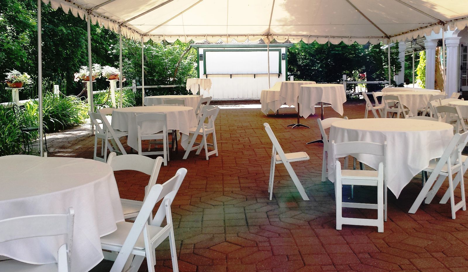A tented outdoor area with round tables and white chairs, surrounded by greenery.