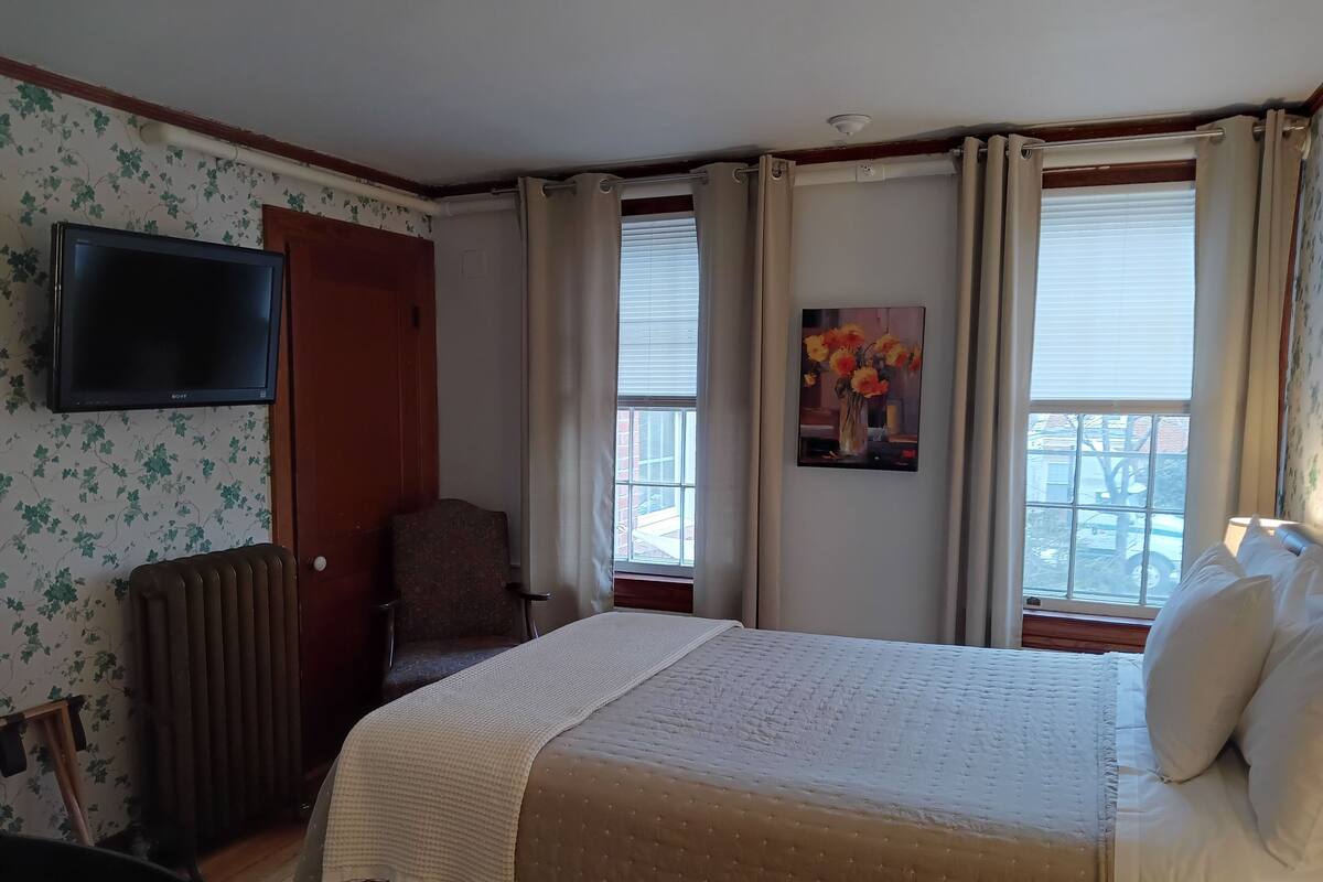 A hotel room with a bed, a chair, a radiator, a television, and a window with curtains.