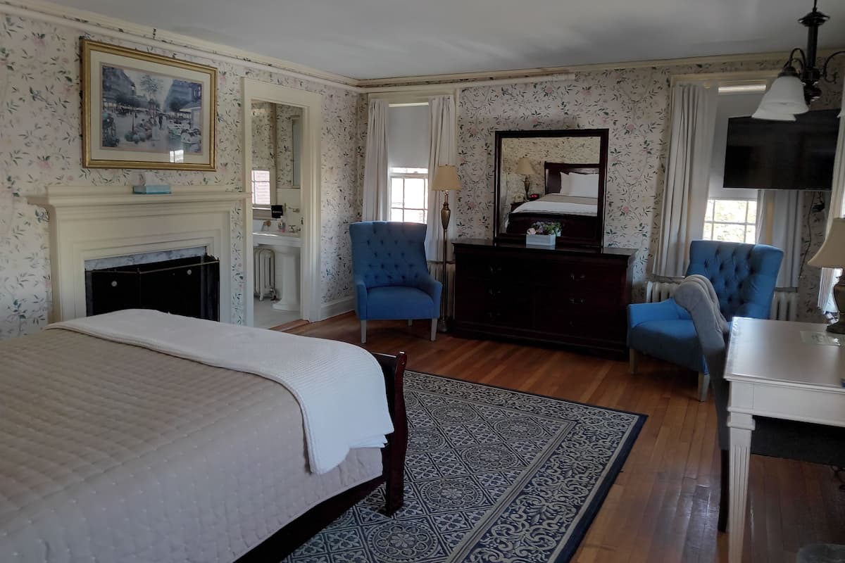 A bedroom with a bed, two blue armchairs, a dresser, a mirror, a fireplace, and a television.