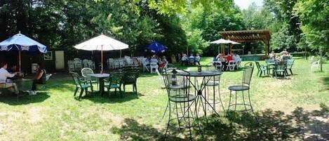 An outdoor seating area with tables and chairs, surrounded by trees and covered by umbrellas.