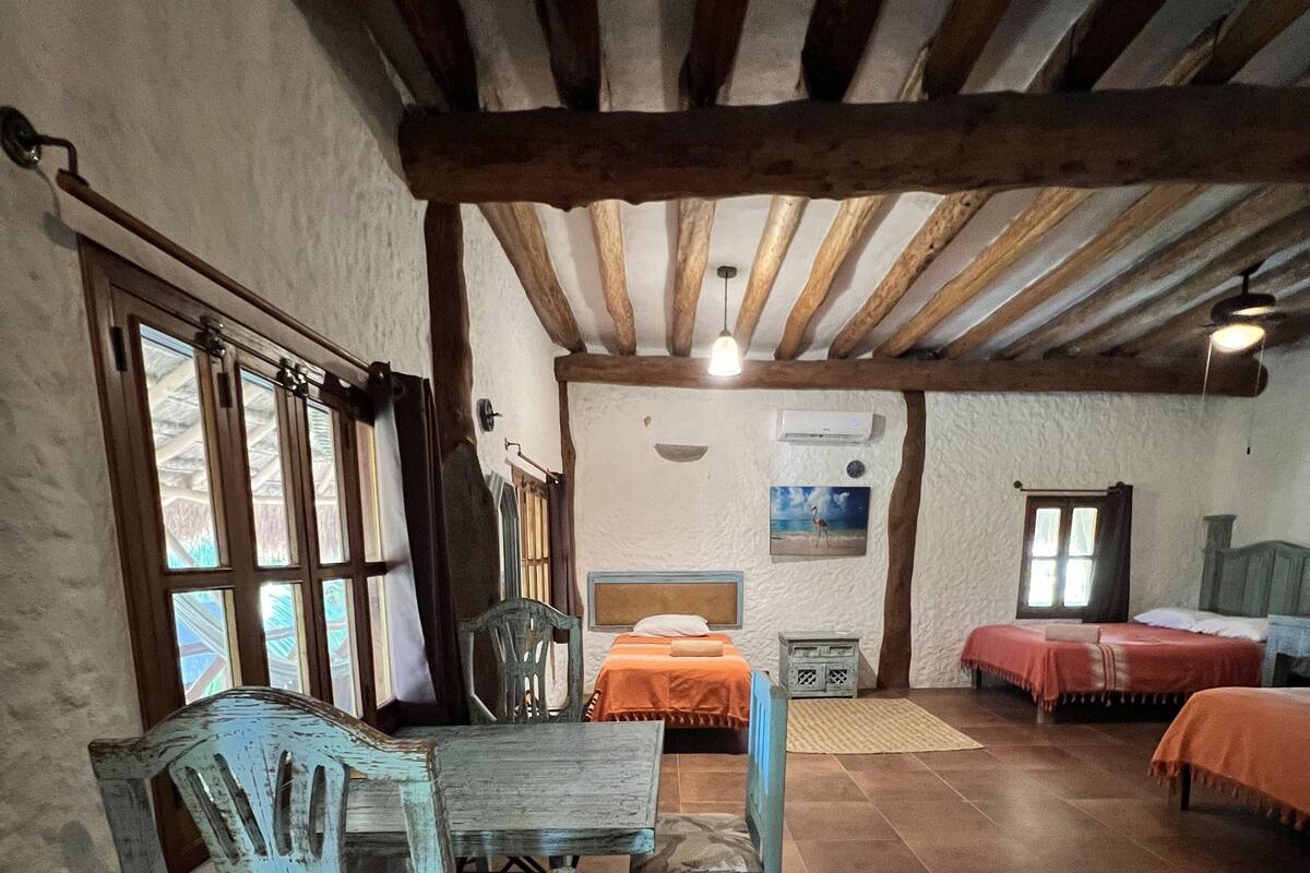 A rustic room with a wooden ceiling, a bed with an orange bedspread, a wooden table with chairs, and a window with curtains.