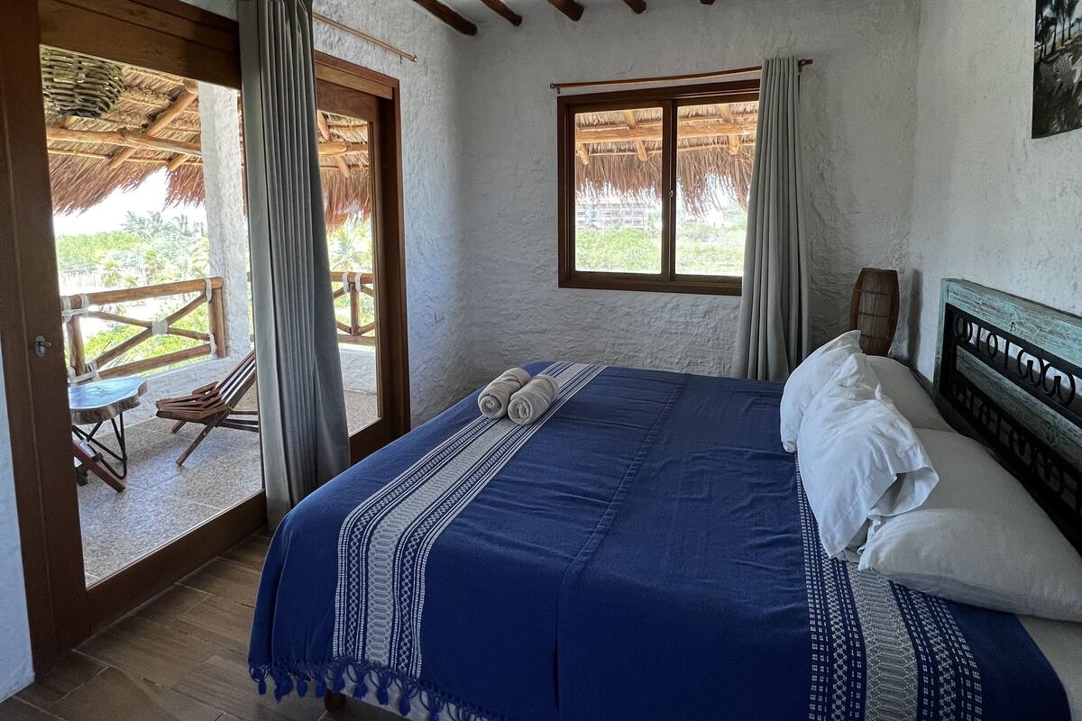 A bedroom with a bed, wooden beams, a ceiling fan, and a view of the outdoors.