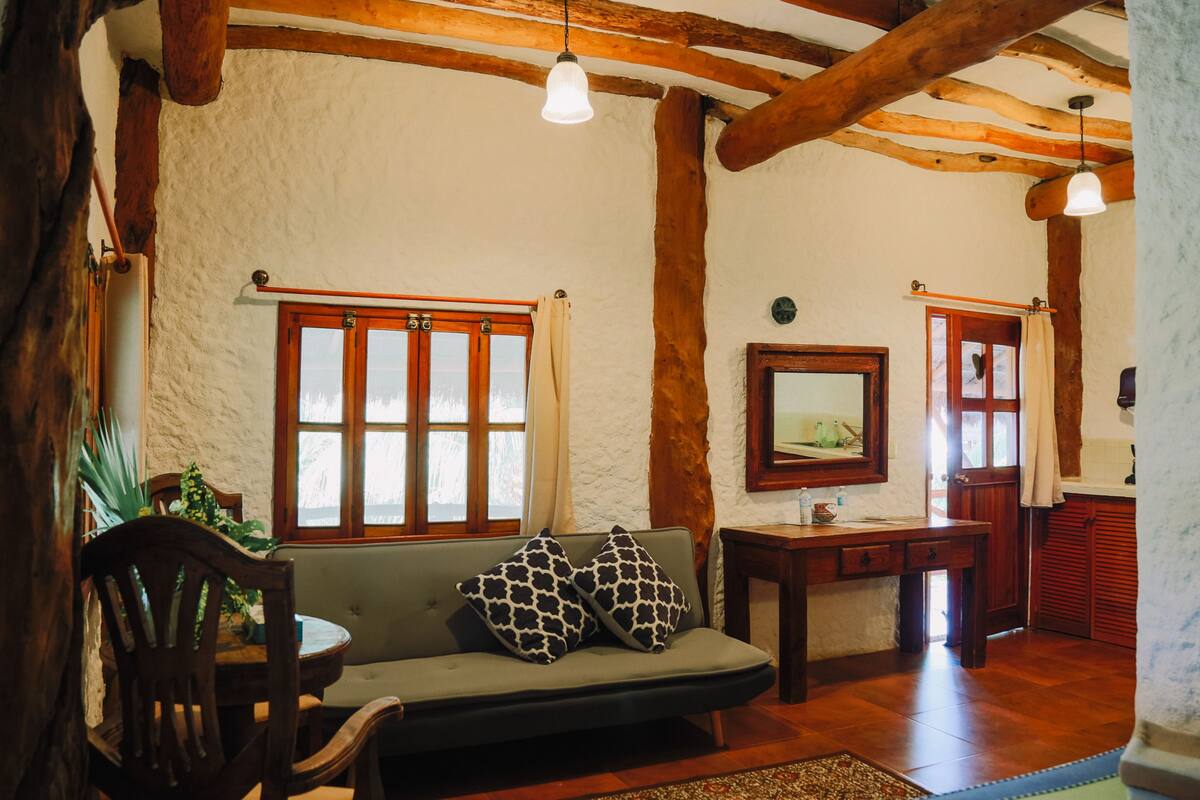 A cozy living room with a sofa, wooden beams, a dining table, and a window with curtains.