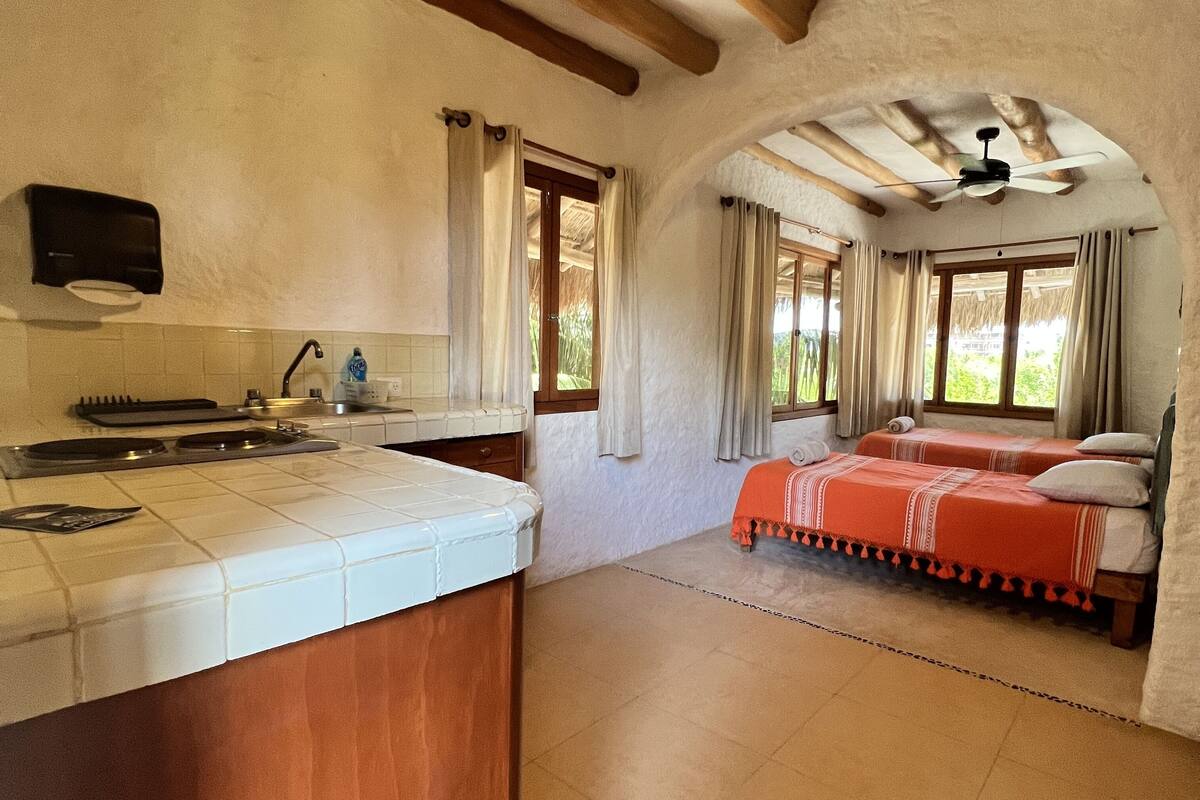 A room with a kitchen area, two beds with red bedding, a ceiling fan, and a view of greenery outside.